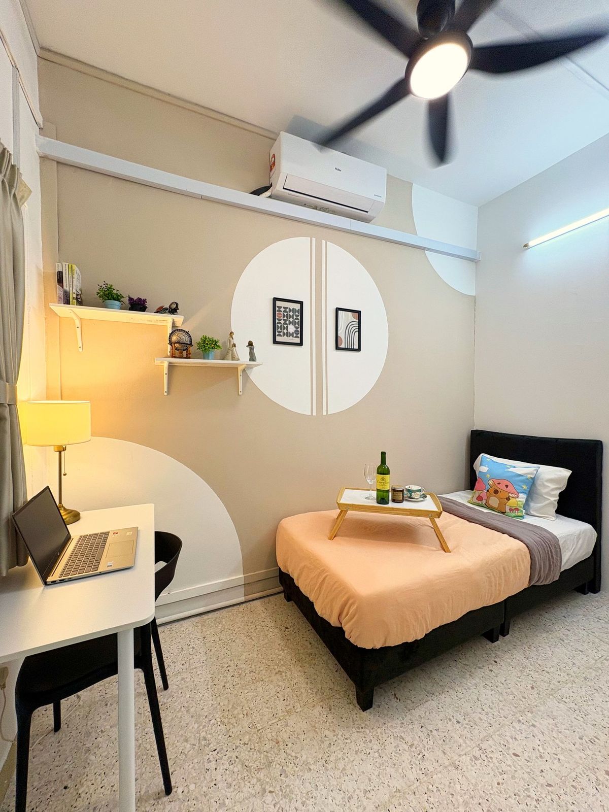 Bedroom with a peach single bed, breakfast tray, white study desk with a laptop and lamp, and circular wall art