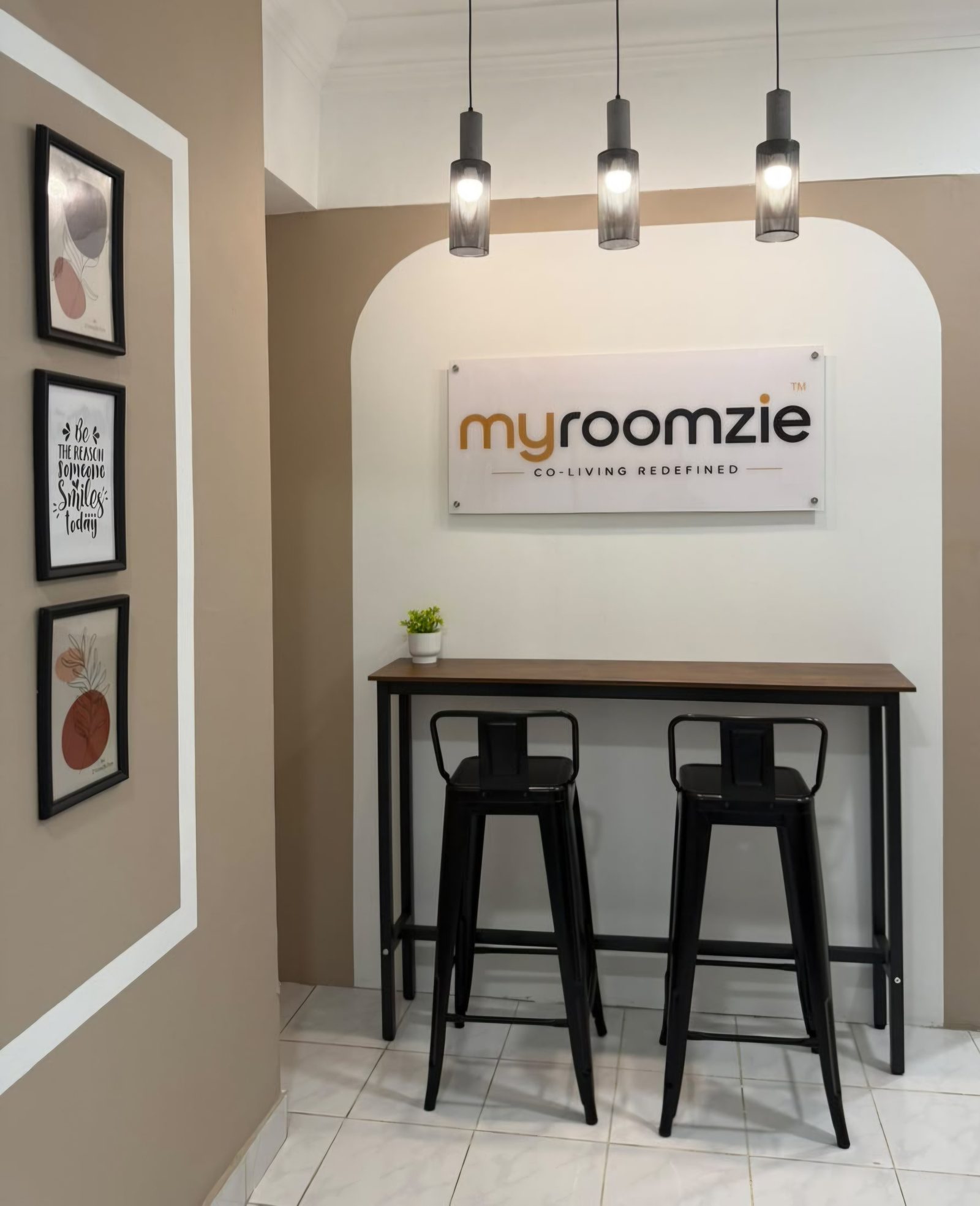 Myroomzie sign above a wooden bar table with two stools, pendant lights, and framed prints